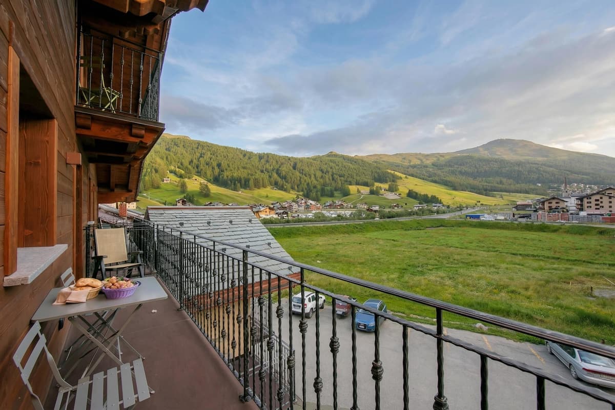 A balcony with a table, chairs, and a view of a green valley and distant buildings.