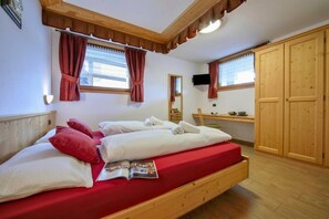 A wooden bed with red bedding, a wooden desk, a television, and a wardrobe in a room with wooden floors and white walls.
