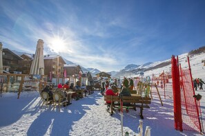 A snow-covered outdoor seating area with wooden benches and tables, surrounded by buildings and a ski slope in the background.