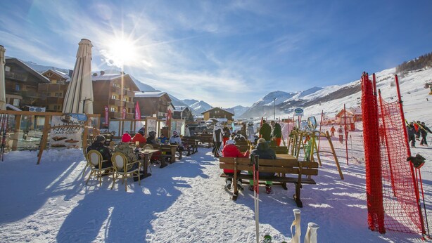 A snow-covered outdoor seating area with wooden benches and tables, surrounded by buildings and a ski slope in the background. - Tamigi (Livigno)