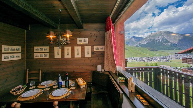Une table de salle à manger en bois, dressée avec des assiettes, des verres et des couverts, et offrant une vue sur la montagne à travers la rambarde d’un balcon.