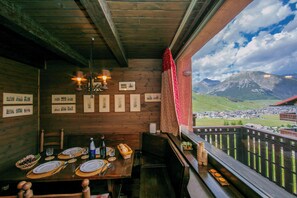 A wooden dining table set with plates, glasses, and cutlery, with a mountain view through a balcony railing. - Arianna (Livigno)