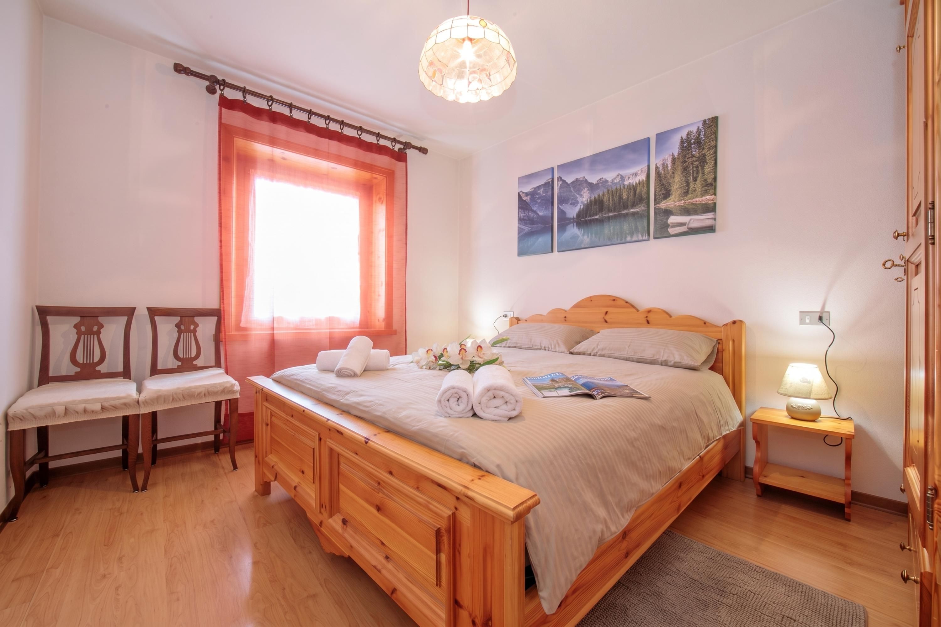 A bedroom with a wooden bed, a window with curtains, a desk with a lamp, and a wall-mounted picture.