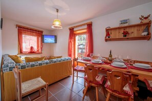 A cozy living room with a sofa, dining table set for two, a television, and a wooden shelf with decorations.