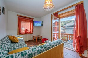A room with a sofa, a television, a wooden table, and a balcony with a view of a building and trees. - Martina Ski in - Ski out Mt.50 (Livigno)