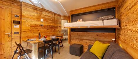 A compact room with a bunk bed, a dining table with chairs, a sofa, and a small kitchenette.