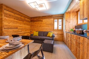 Una habitación acogedora con paredes de madera, una mesa de comedor preparada para dos personas, un sofá con cojines y una zona de cocina con estufa.