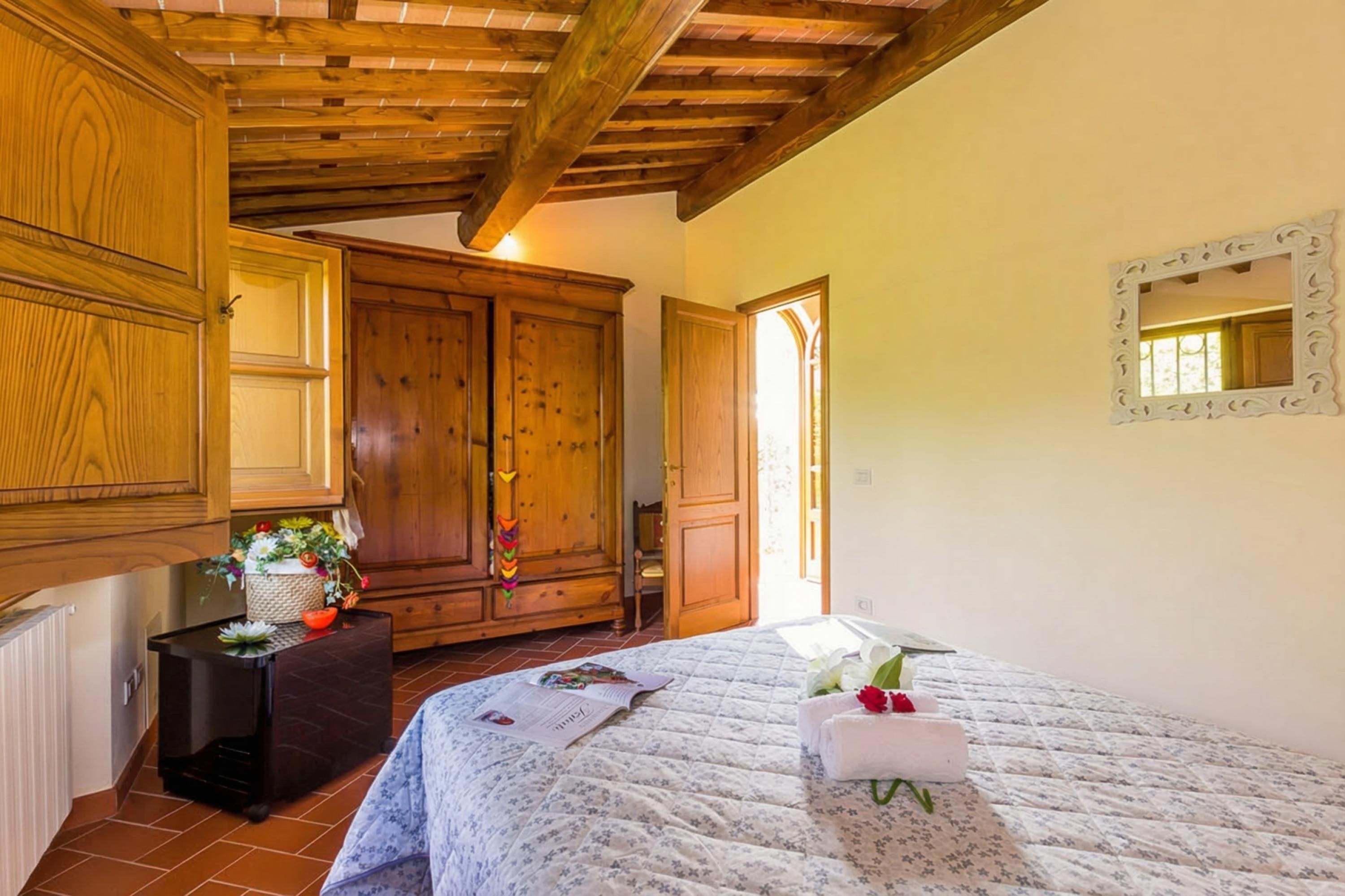 A bedroom with a wooden wardrobe, a bedside table with a vase of flowers, a bed with a floral bedspread, and a mirror on the wall.