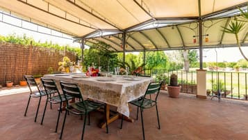 An outdoor patio with a table set for dining, surrounded by chairs, under a covered area with a view of greenery.