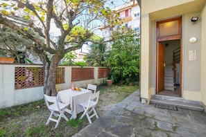 Pátio com mesa, cadeiras, uma árvore e entrada de um edifício.