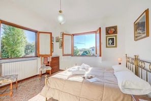 Quarto com uma janela ampla, uma cama com roupa branca, uma cômoda de madeira e uma cadeira.