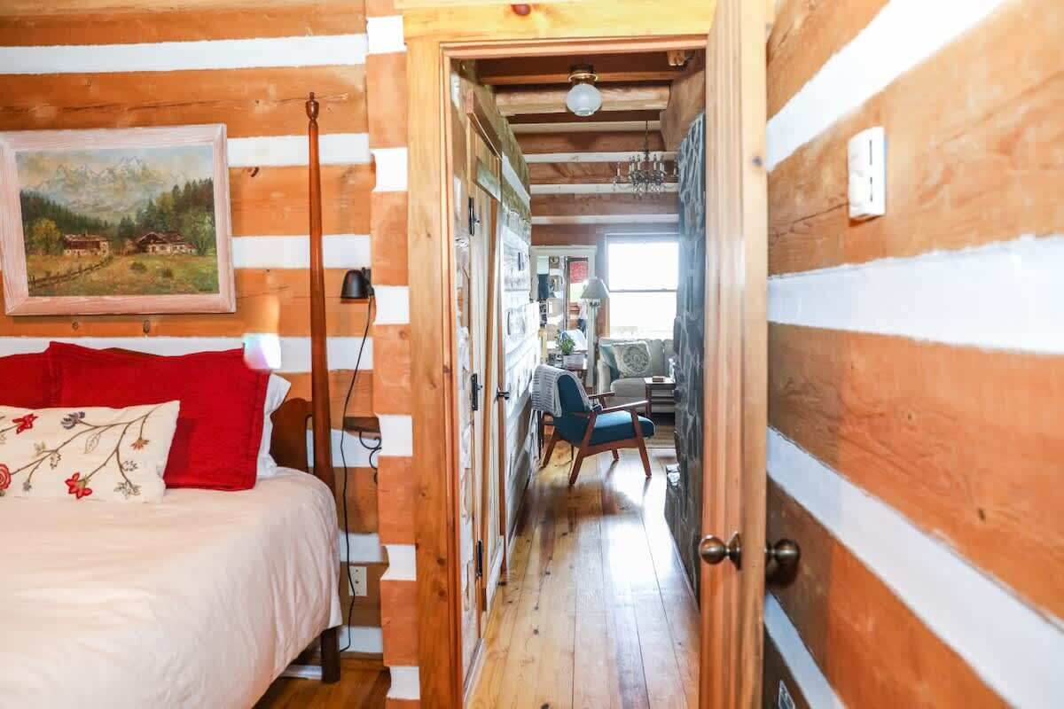 A wooden interior with a bed, a chair, and a hallway leading to a room with a window.