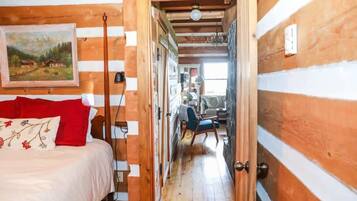 A wooden interior with a bed, a chair, and a hallway leading to a room with a window.