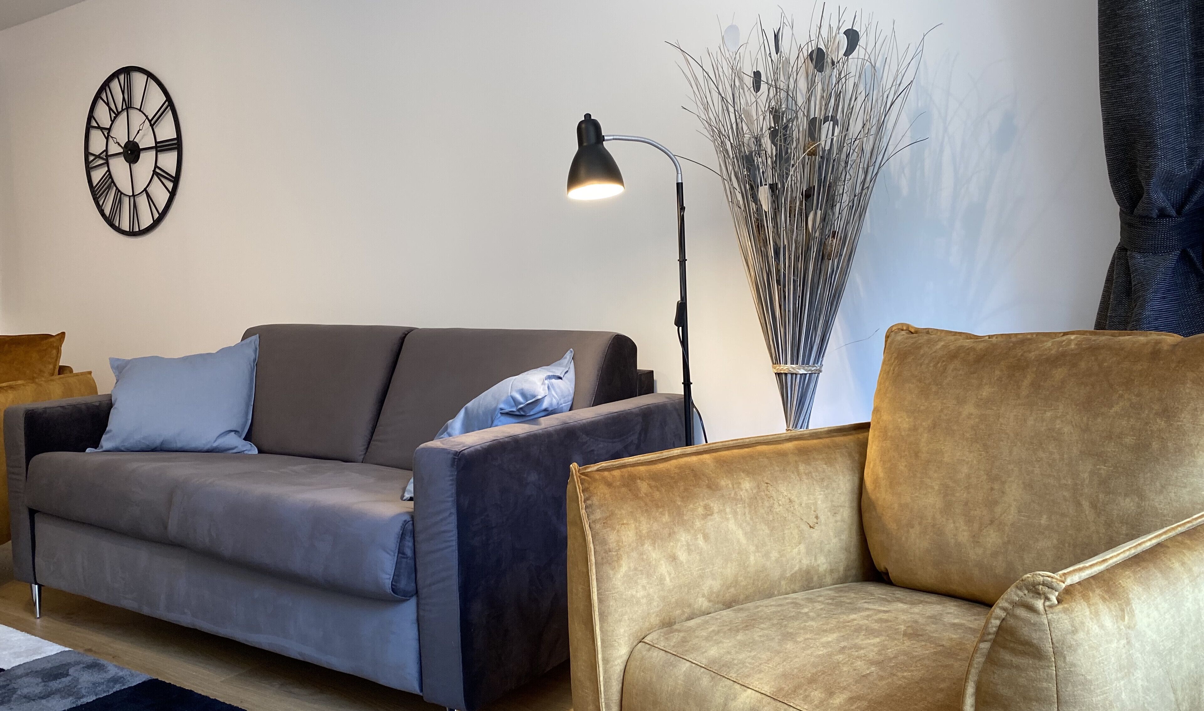 A living room with a grey sofa, a brown armchair, a floor lamp, and a wall clock.