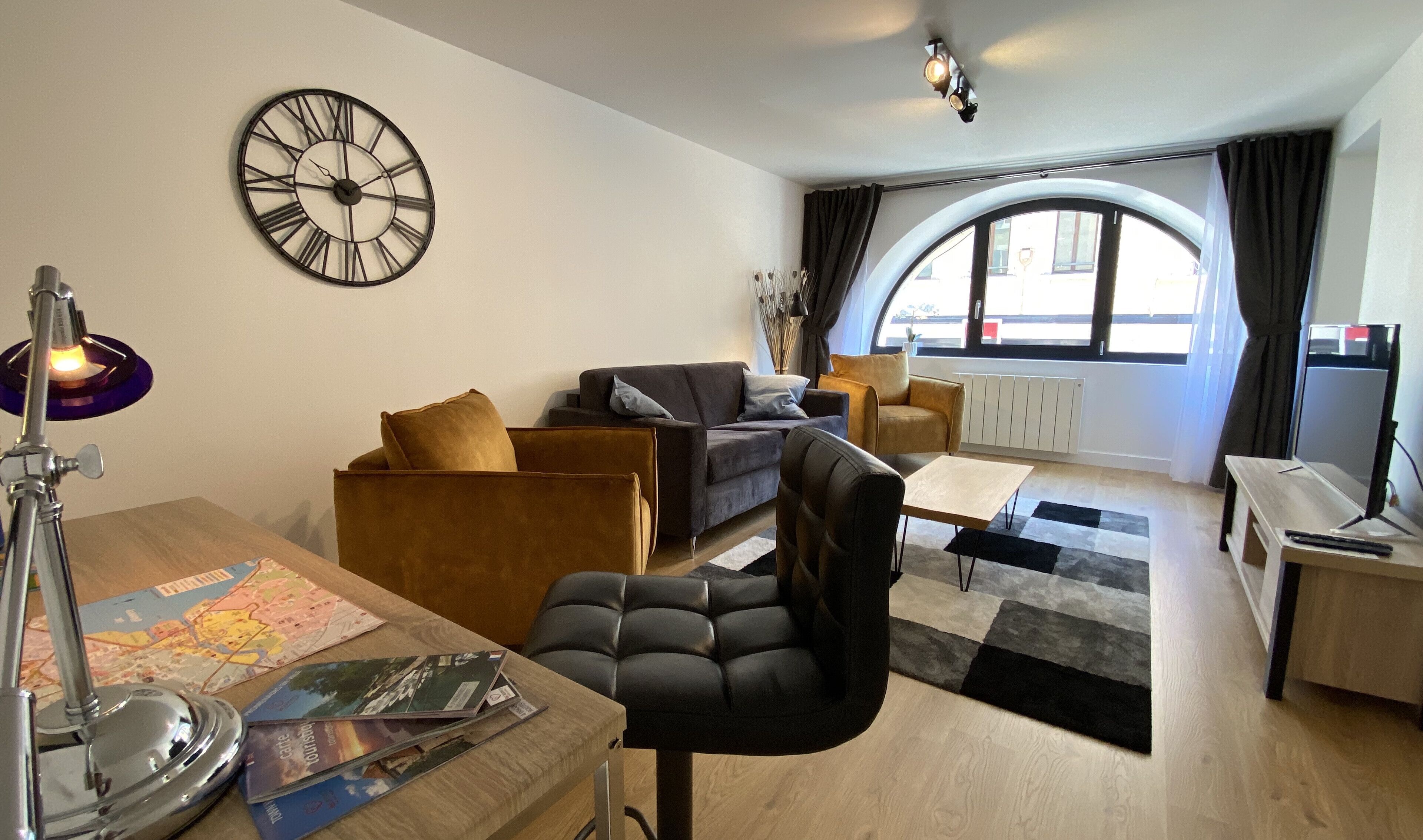 Ein modernes Wohnzimmer mit einer Couch, Sesseln, einem Couchtisch, einem Schreibtisch mit Lampe und einer großen Wanduhr.