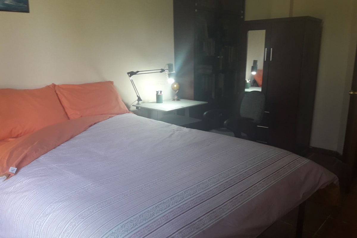 Una cama bien hecha con un edredón estampado, una almohada naranja, una lámpara de noche y un armario oscuro.