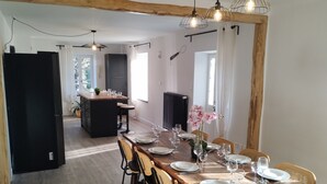 Un comedor con una mesa de madera dispuesta para seis personas, rodeada de sillas. Se ve una cocina con un refrigerador negro al fondo.