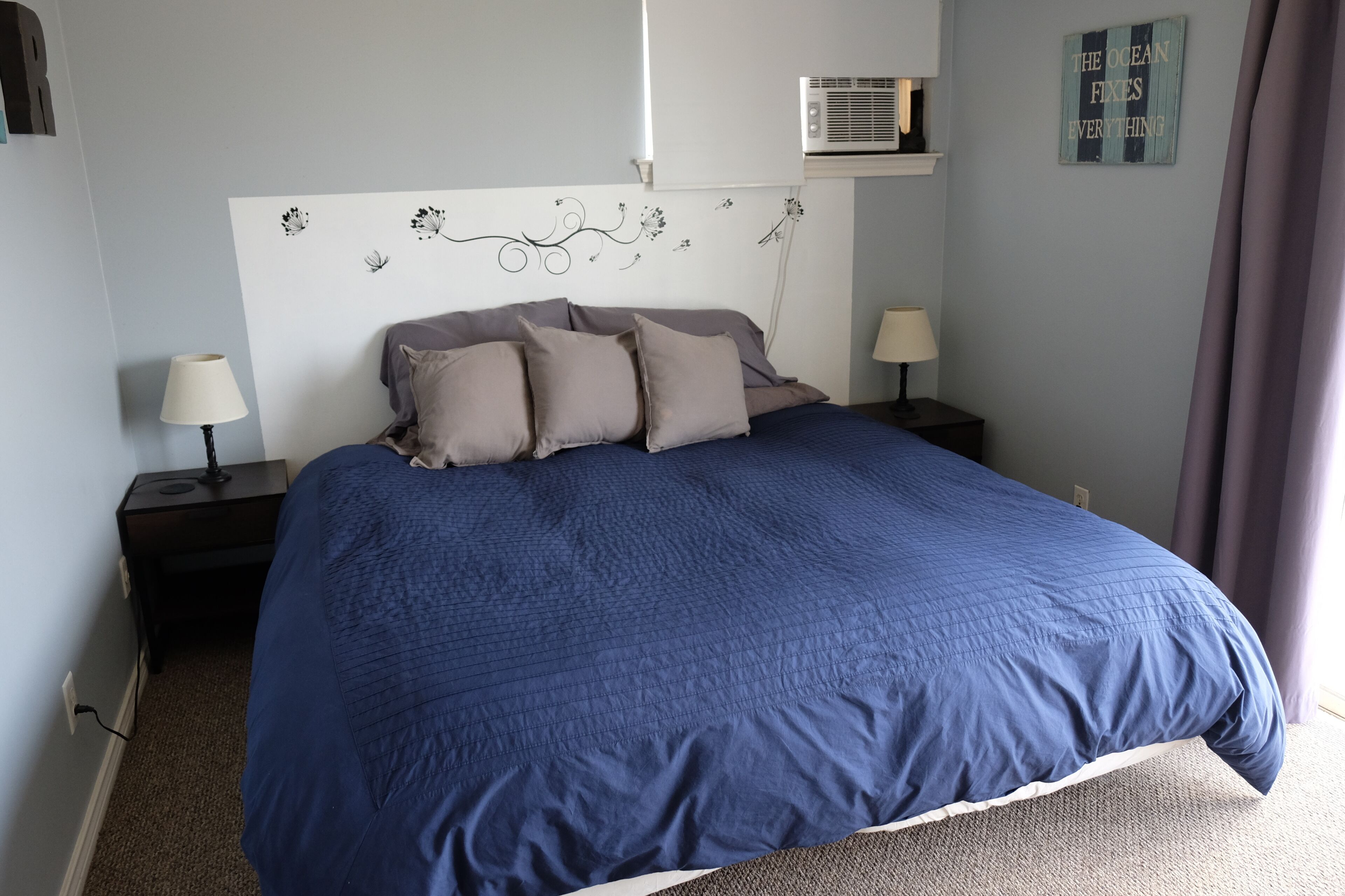 A bedroom with a large bed, two bedside tables with lamps, a wall-mounted air conditioner, and a decorative sign.
