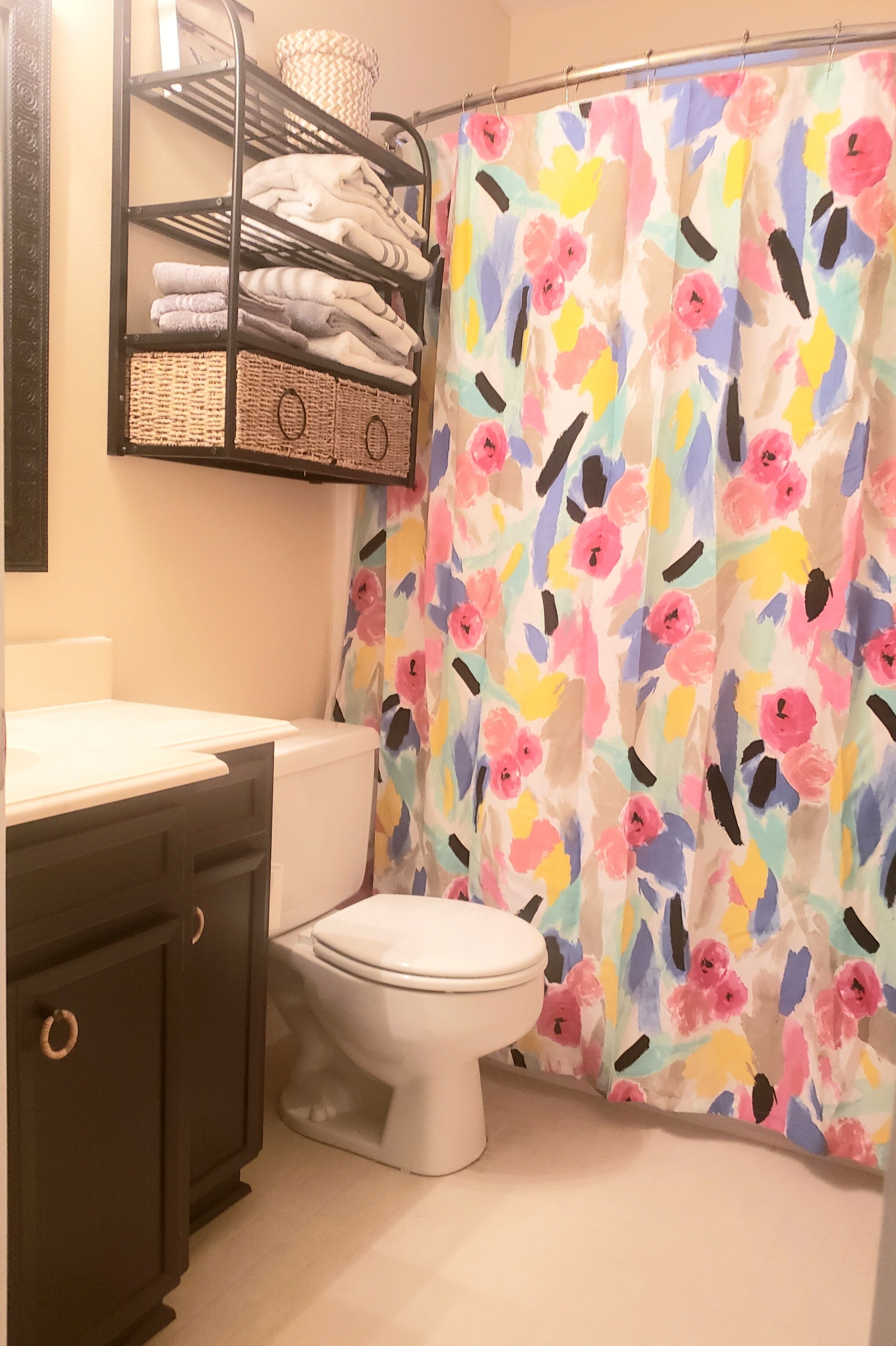 Une salle de bain avec des toilettes blanches, un meuble en bois foncé, une étagère en métal avec des serviettes pliées et un rideau de douche à motifs floraux.
