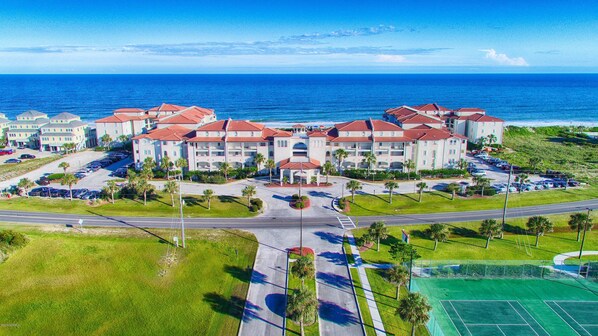 Un hotel frente al mar con varias construcciones, estacionamiento y cancha de tenis.