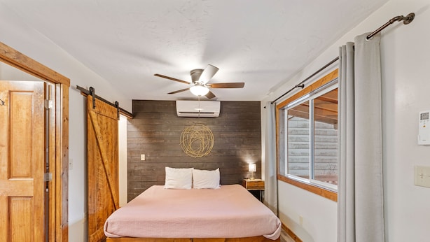 A bedroom with a bed, a ceiling fan, an air conditioner, a wooden floor, and a window with curtains.