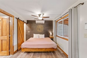 Una camera da letto con un letto, una ventola a soffitto, un condizionatore, pavimento in legno e una finestra con tende.