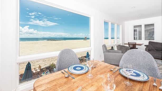 A dining area with a view of the beach, a table set with plates and glasses, and a grey sofa in the background. - Panoramic Oceanfront Views from Beautiful Upper Level Home with Patio (Newport Beach)