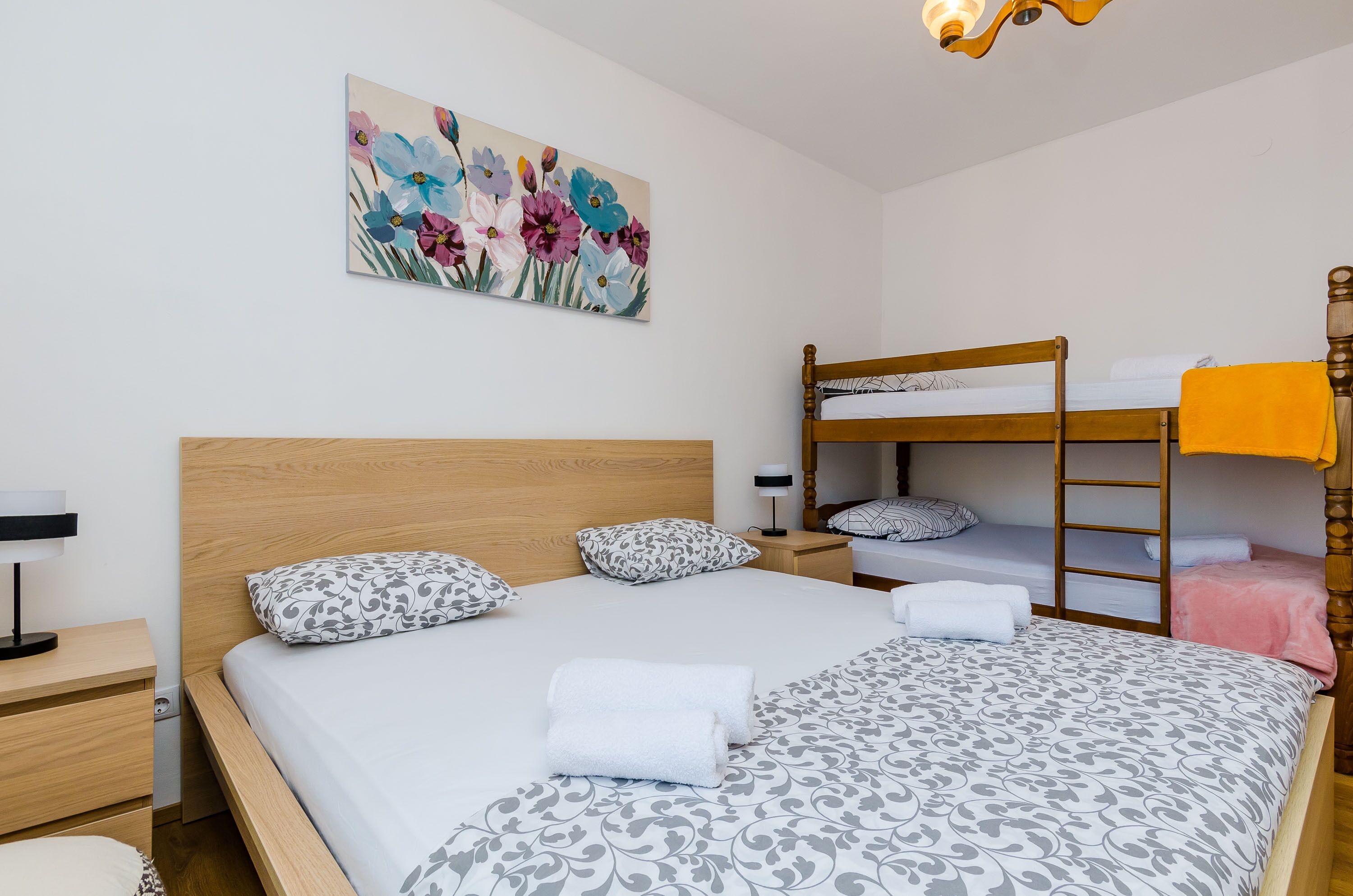 A bedroom with a wooden bed, a bunk bed, a bedside table with a lamp, and a floral painting on the wall.