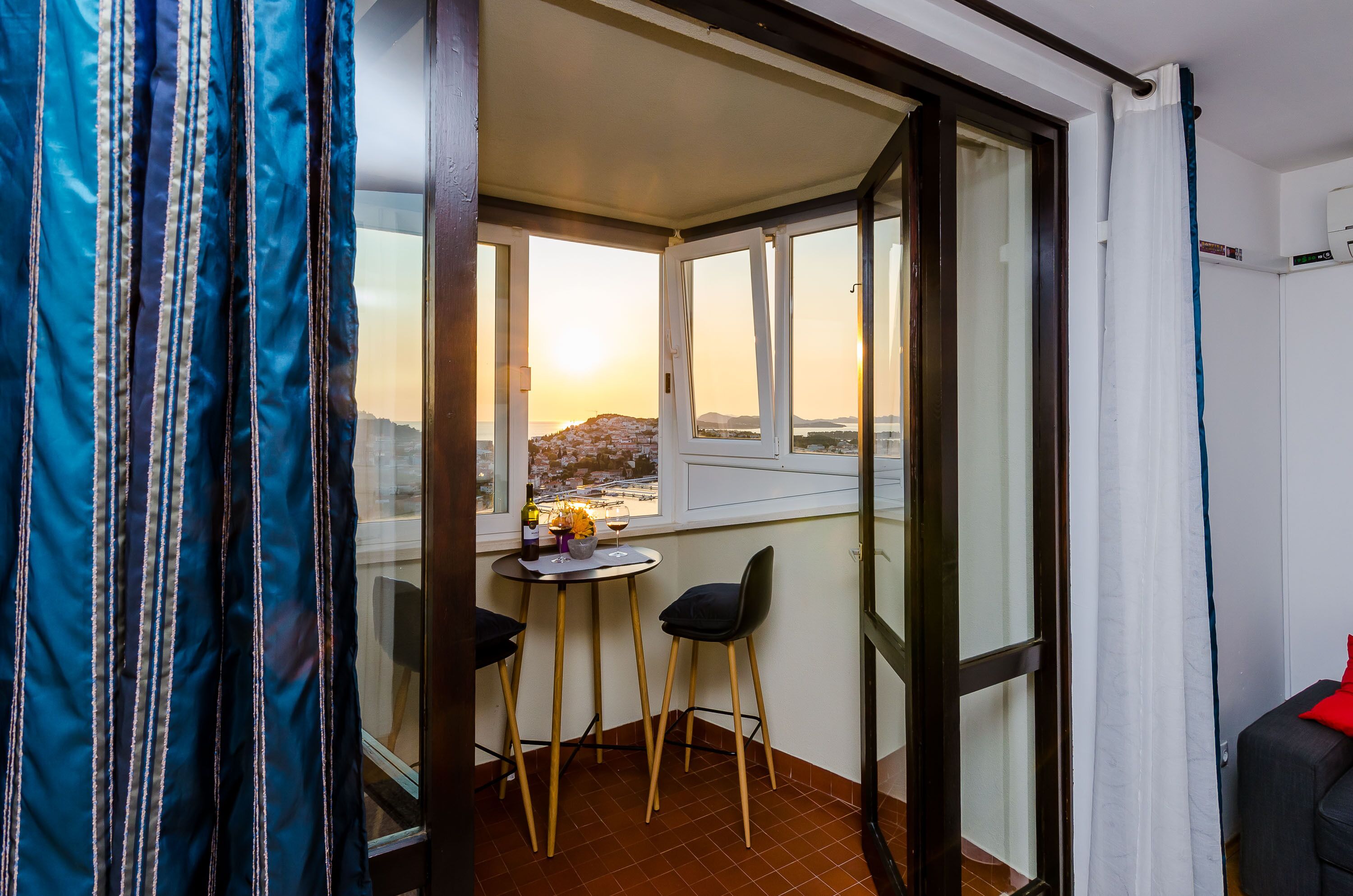 A balcony with a table and chairs, a bottle of wine, and a view of a cityscape at sunset.