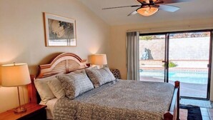 Un dormitorio con una cama grande, dos mesitas de noche con lámparas, un ventilador de techo, un cuadro en la pared y vistas a una piscina exterior.