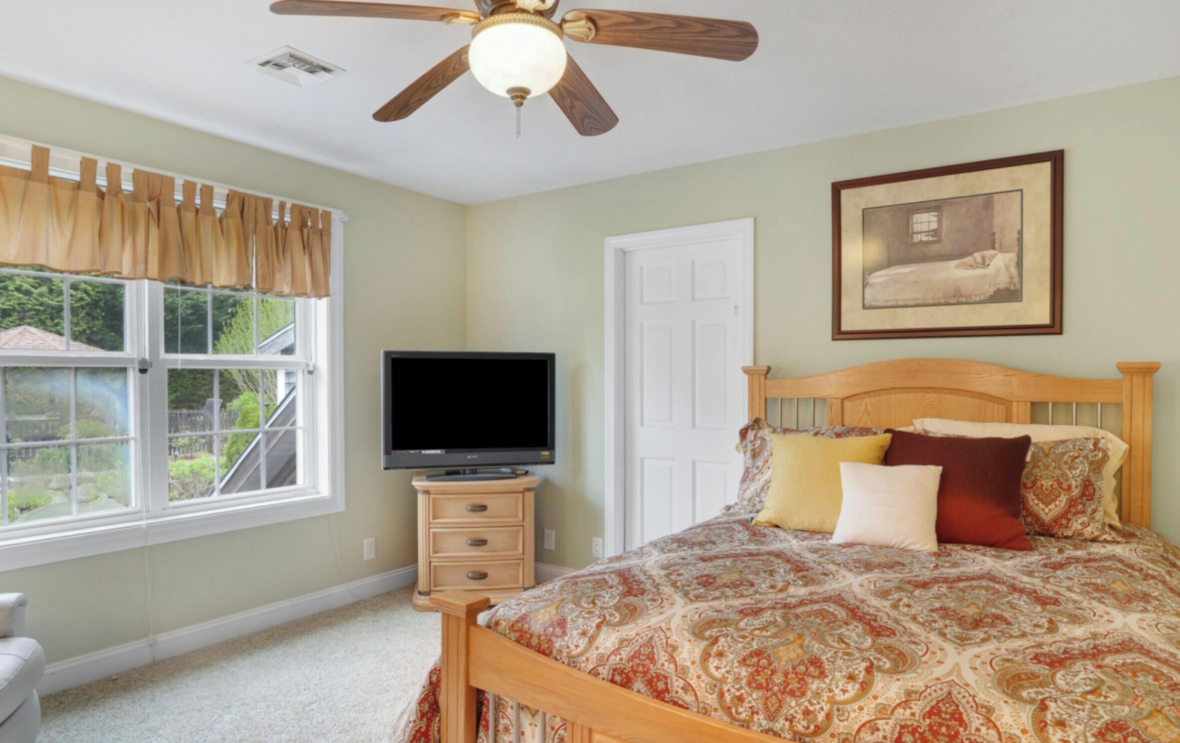 A bedroom with a bed, a ceiling fan, a television, a dresser, and a window with curtains.