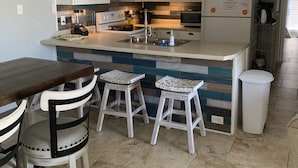 A kitchen with white cabinets, a built-in oven, a microwave, and a refrigerator. There are two stools and a wooden table.