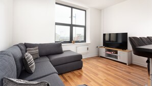 Een moderne woonkamer met een grijze hoekbank, een flatscreen televisie op een houten dressoir, en een groot raam met uitzicht op de stad.