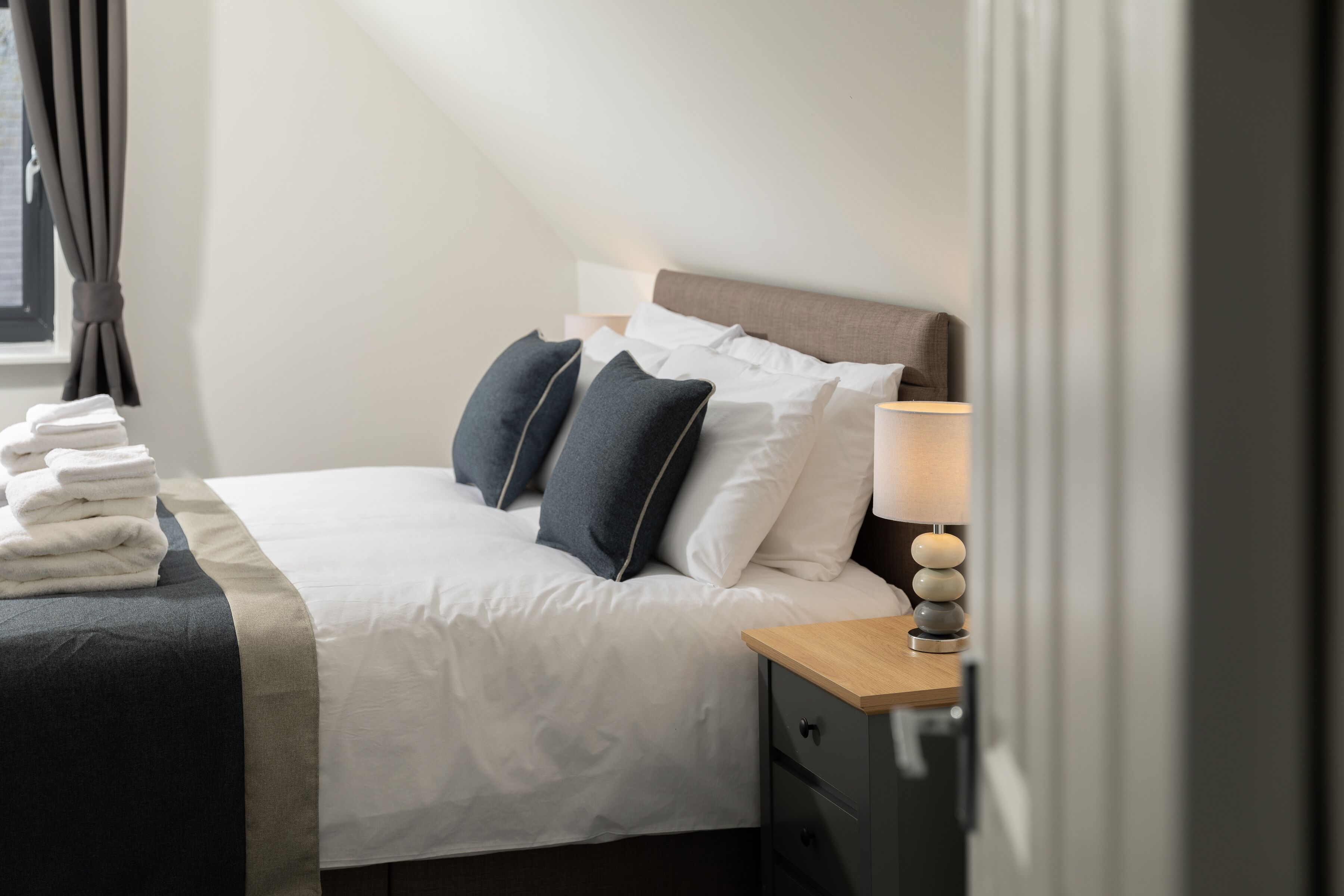 A neatly made bed with a white duvet, two dark pillows, and a wooden nightstand with a lamp.