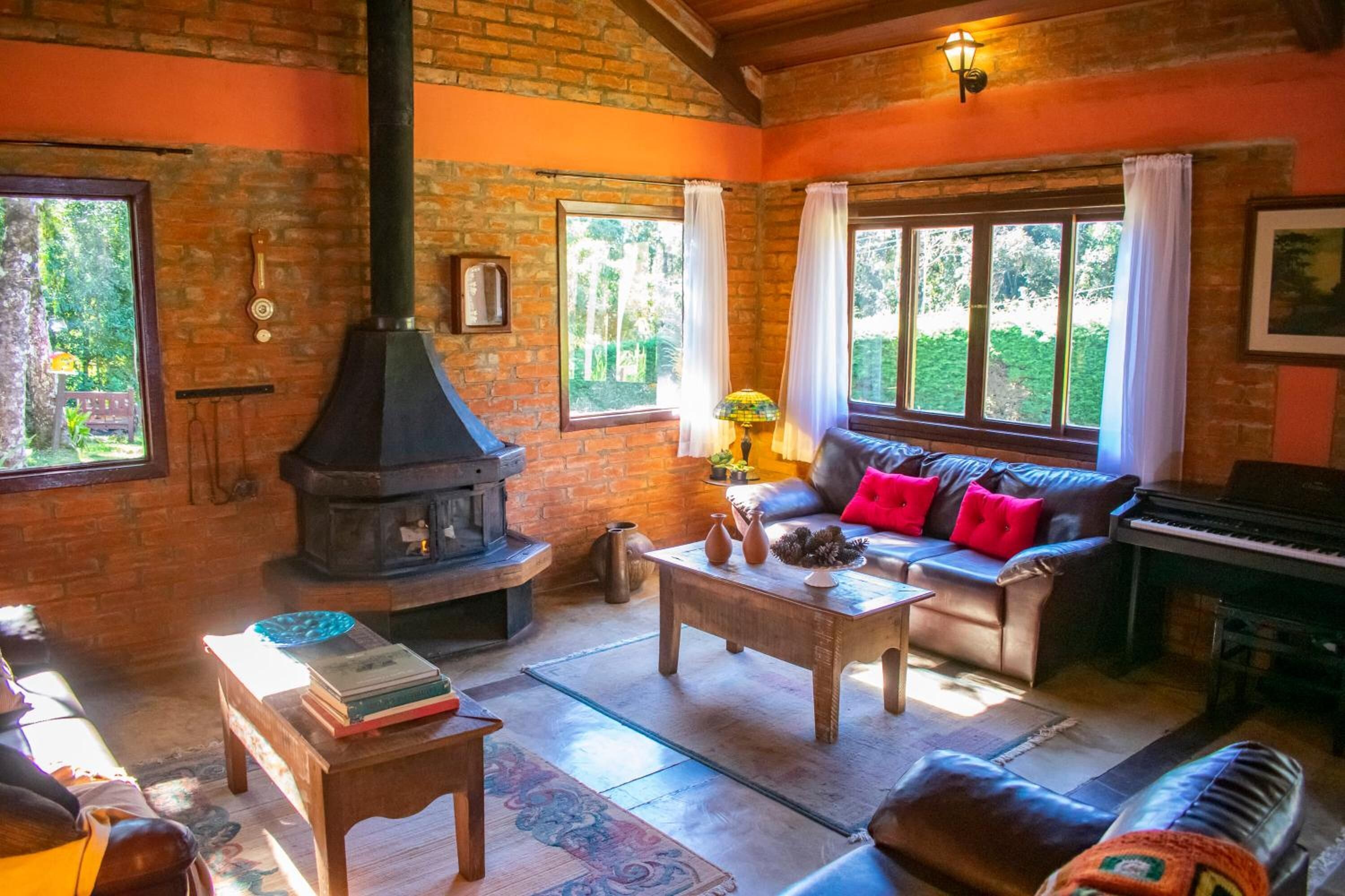 A cozy living room with a brick fireplace, a sofa with red cushions, a piano, and a large window with curtains.