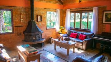 A cozy living room with a brick fireplace, a sofa with red cushions, a piano, and a large window with curtains.