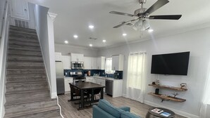 A modern kitchen with white cabinets, a dining area, a living room with blue sofas, and a staircase.