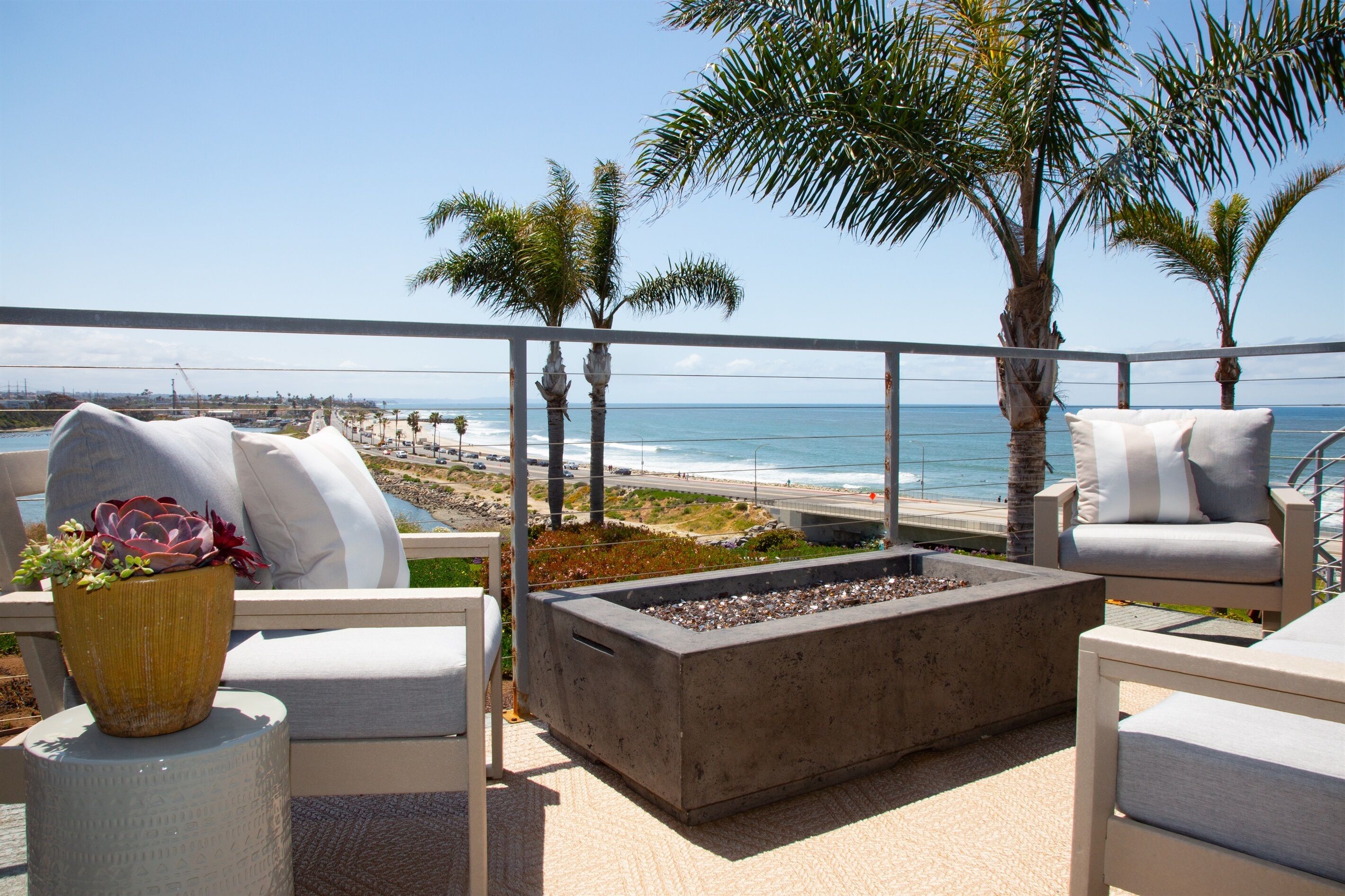 A balcony with a view of the beach, outdoor seating, and a fire pit.