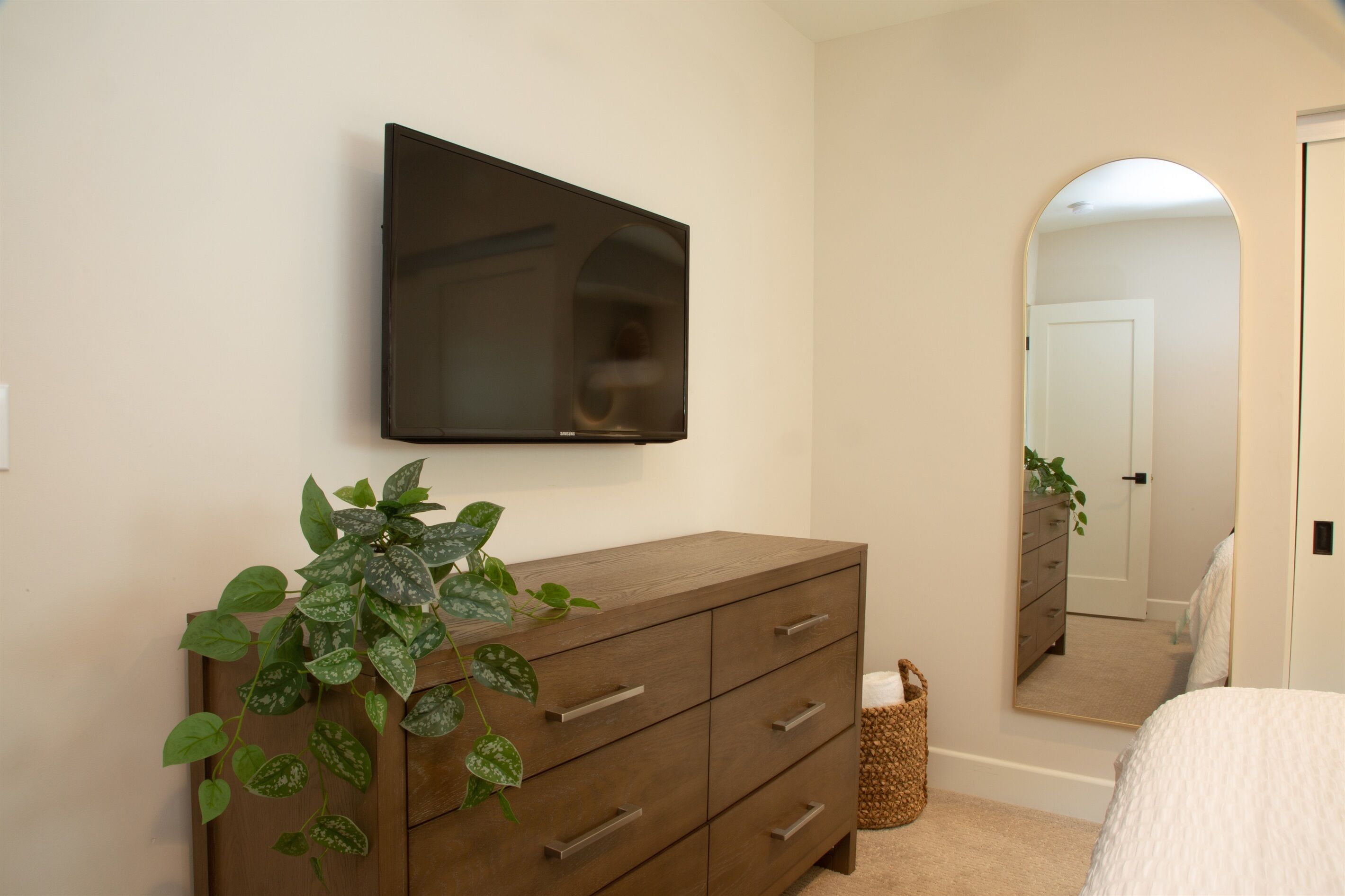 A bedroom with a wooden dresser, a flat-screen TV mounted on the wall, a mirror, and a potted plant.