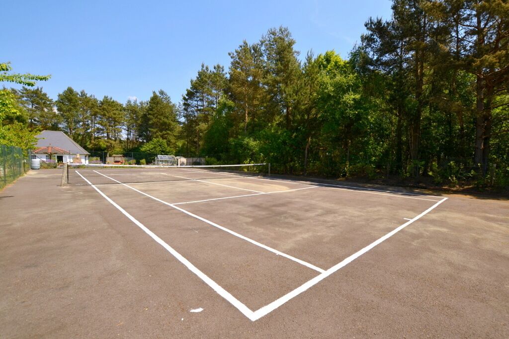 Un campo da tennis all'aperto con una rete, circondato da alberi e un edificio sullo sfondo.