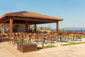 Un área con mesas y sillas de madera, un pabellón con techo de paja y vista al mar.