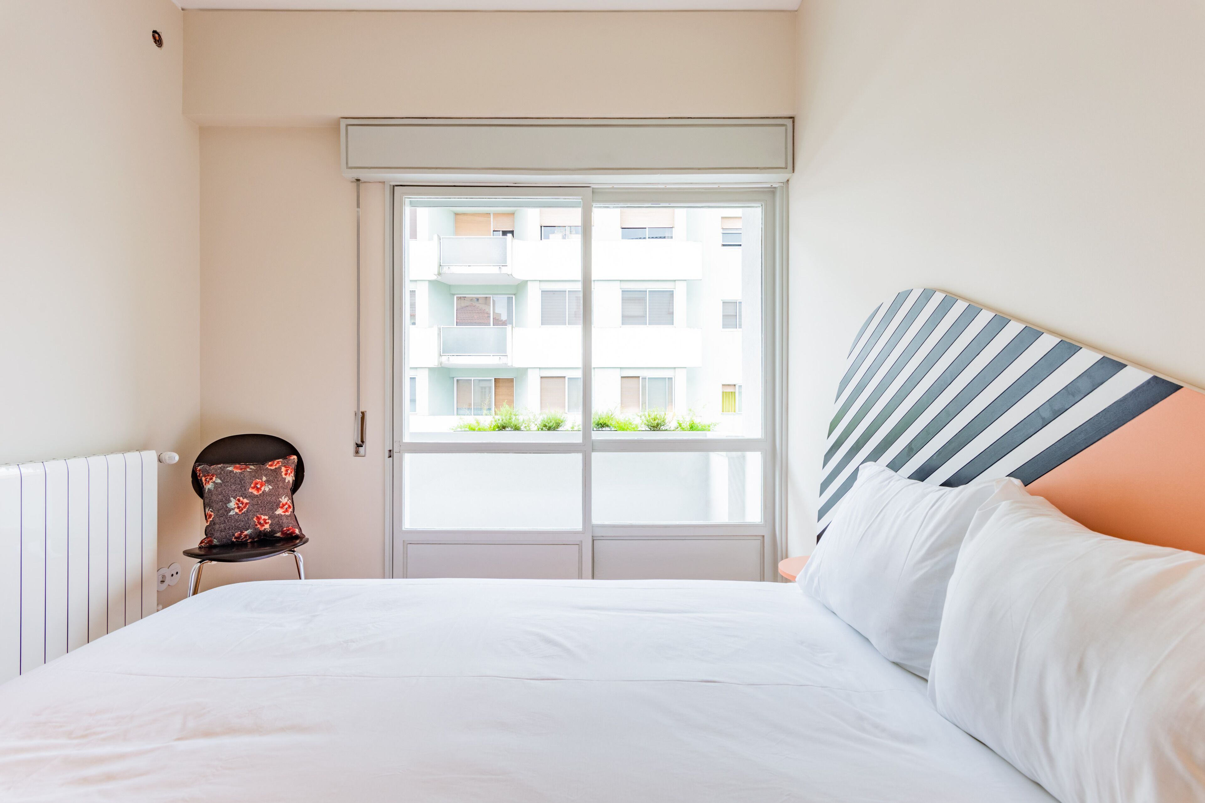 Une chambre d’hôtel avec un lit, une chaise dotée d’un coussin à motifs floraux, une fenêtre donnant sur des immeubles et une tête de lit à rayures.