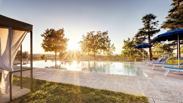 Une piscine entourée d’arbres, avec des chaises longues et des parasols, sous un ciel dégagé.