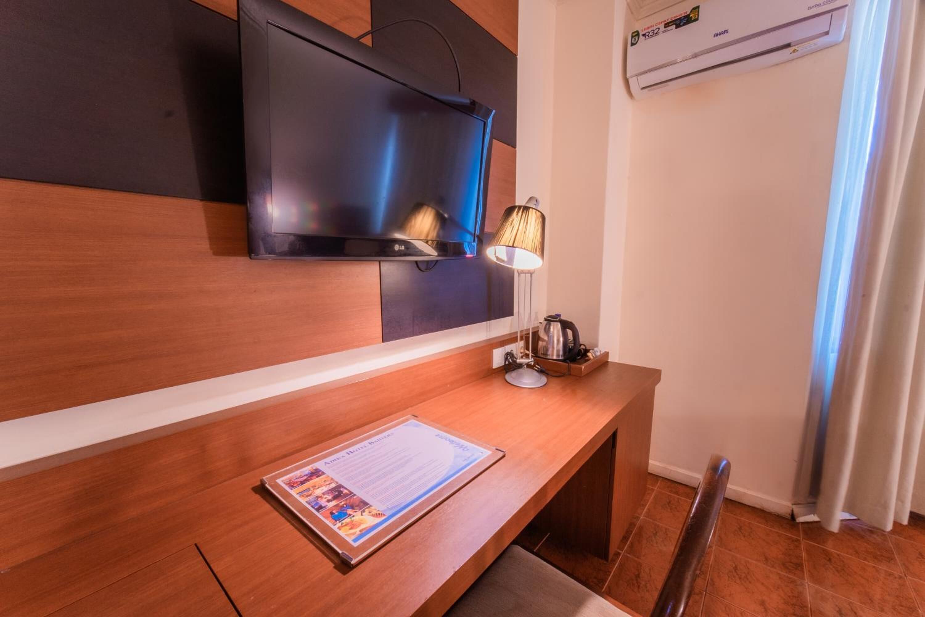 A hotel room with a wooden desk, a flat-screen TV mounted on the wall, a lamp, and a kettle.