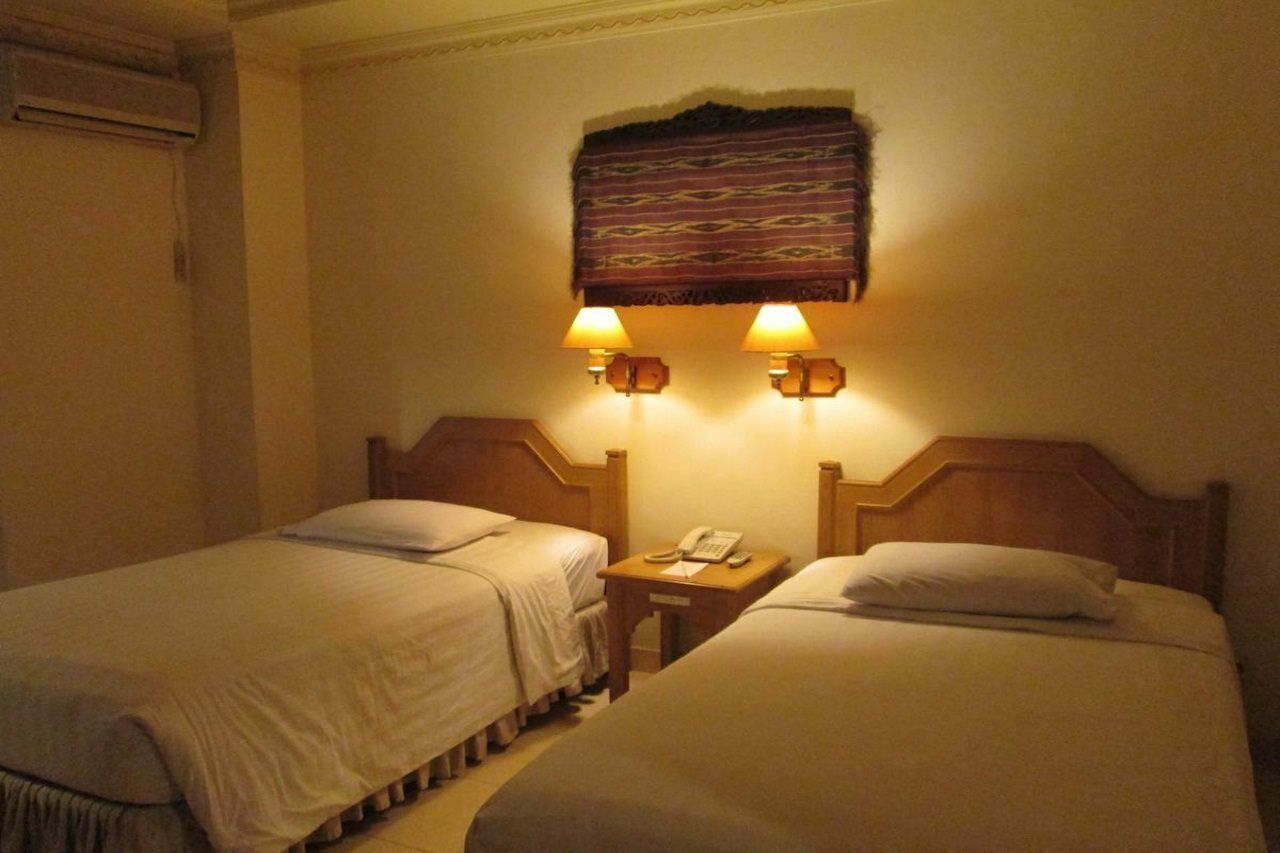 A hotel room with two single beds, a small bedside table with a phone, and a wall-mounted lamp.