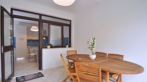 A dining area with a wooden table and chairs, a potted plant, and a view of a kitchen through a glass door. - Appartamento a Capaccio ID 3338 (Licinella-Torre di Paestum)