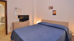 A bedroom with a bed, a TV on a wooden dresser, and a nightstand with a lamp. - Appartamento a Capaccio ID 3338 (Licinella-Torre di Paestum)