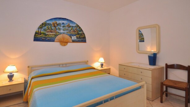 Un dormitorio con cama, dos lámparas de noche, un tocador, una silla, un espejo en la pared y un cuadro de paisaje.
