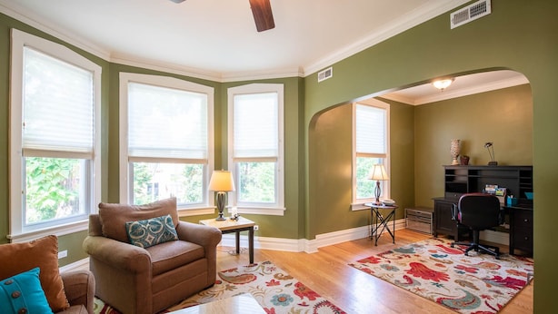 A living room with a brown sofa, a coffee table, and a patterned rug. There are two windows with blinds, a ceiling fan, and a desk with a chair. - Roscoe Village- Only steps to cute shops & dining! Sleeps 4-6 (Chicago)