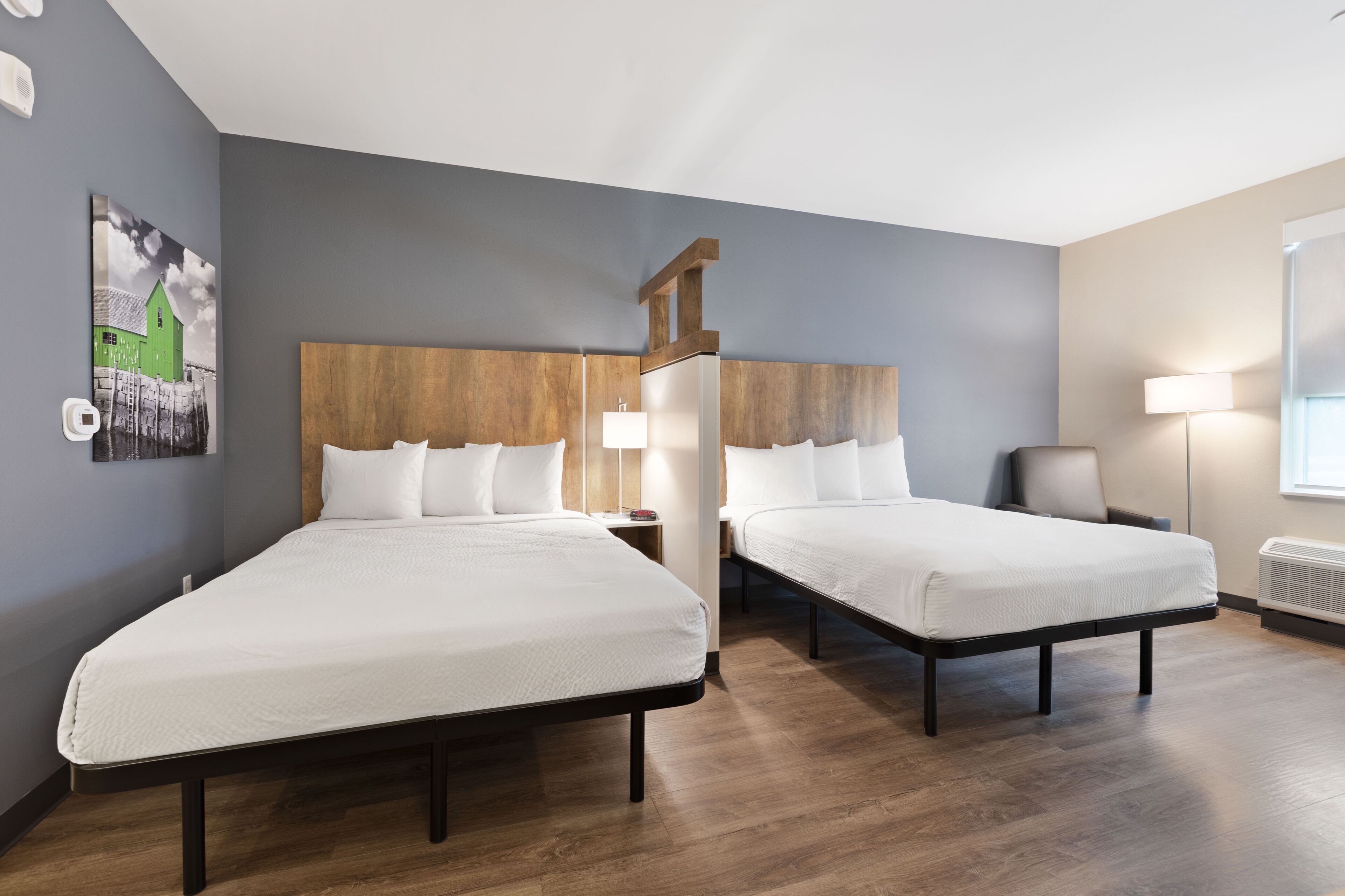 Habitación de hotel con dos camas individuales, cabecera de madera, mesita de noche con lámpara y aire acondicionado instalado en la pared.